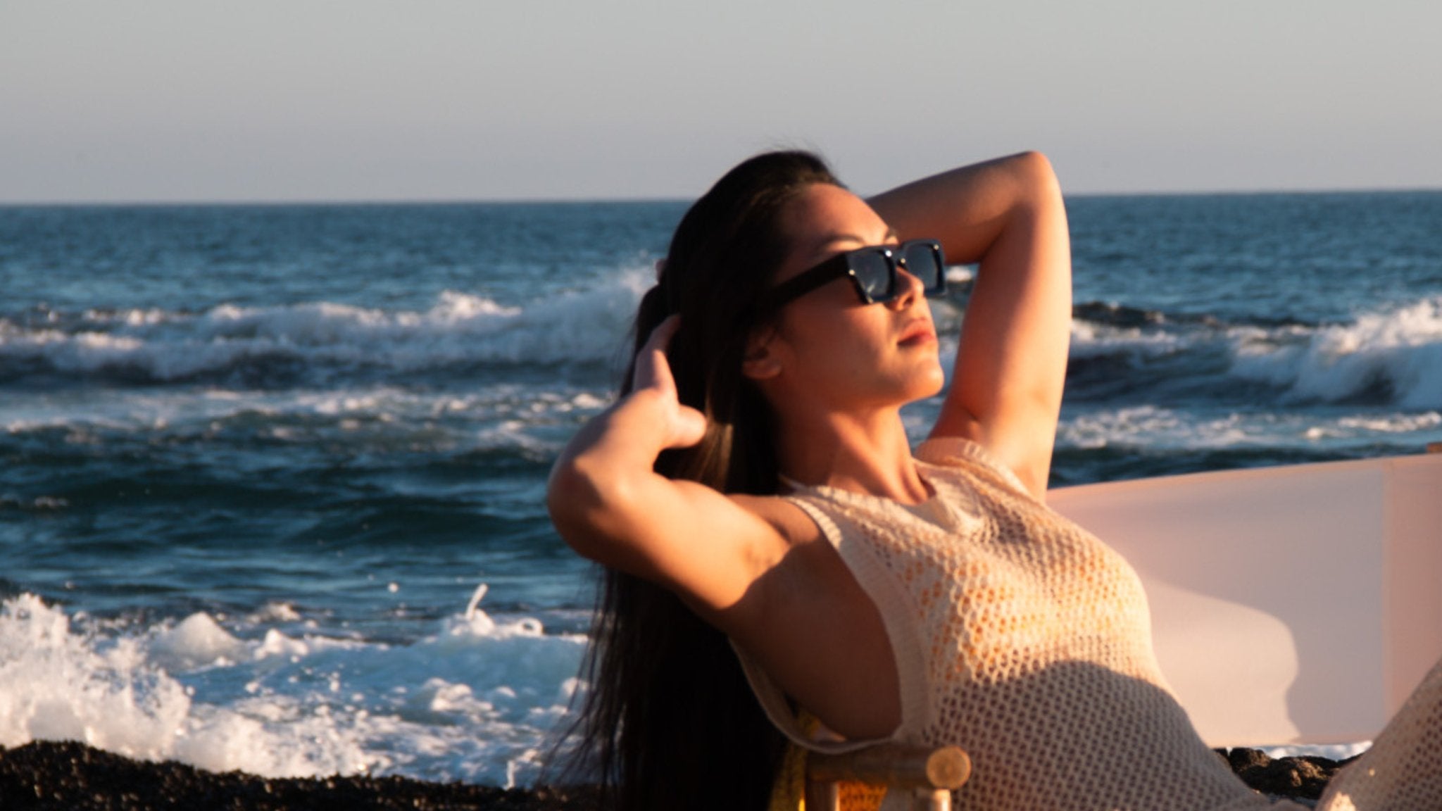 Summer hair care tips hero image – woman relaxing seaside running fingers through glossy hair at golden hour.